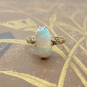 May include: A vintage gold ring featuring an oval opal gemstone. The opal displays a play of color with hints of blue and green. The ring has an ornate, engraved band with decorative detailing on the shoulders.