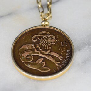 May include: A round, bronze-toned Capricorn zodiac pendant necklace. The pendant features a detailed relief of a goat and the word "CAPRICORN". The necklace has a gold-colored chain and clasp.