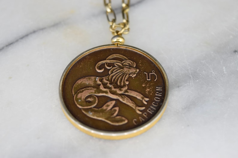 May include: A round, bronze-toned Capricorn zodiac pendant necklace. The pendant features a detailed relief of a goat and the word "CAPRICORN". The necklace has a gold-colored chain and clasp.