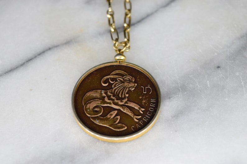 May include: A gold-toned necklace featuring a circular pendant with a Capricorn zodiac symbol. The pendant has a bronze-colored background with a detailed relief of a goat-like creature. The word "CAPRICORN" is inscribed on the pendant.