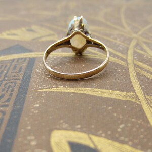 May include: A gold ring with a light-colored oval gemstone. The ring's band is thin and simple, while the setting around the gemstone has intricate detailing. The ring is set against a brown background.