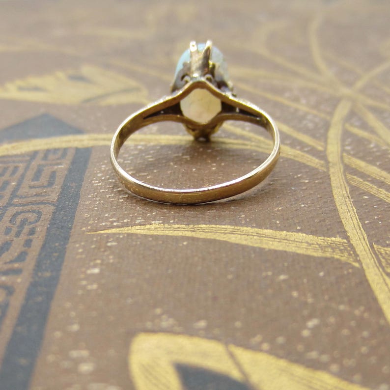May include: A gold ring with a light-colored oval gemstone. The ring's band is thin and simple, while the setting around the gemstone has intricate detailing. The ring is set against a brown background.