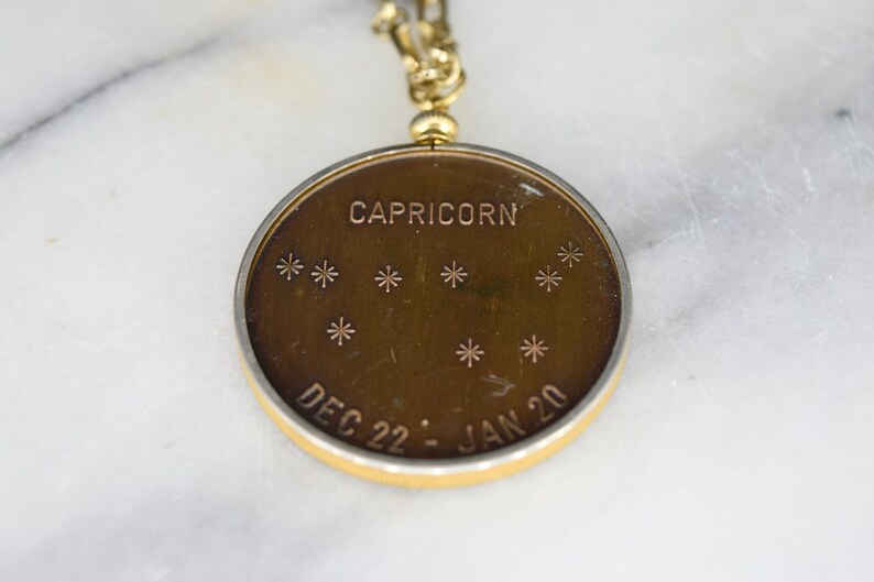 May include: A gold-toned Capricorn pendant with a constellation design. The pendant features the word "CAPRICORN" at the top, with star symbols below. The dates "DEC 22 - JAN 20" are inscribed at the bottom. The pendant is attached to a gold chain.