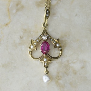 Puede incluir: Un collar de oro con un colgante de rubí y detalles de perlas. El colgante tiene forma de corazón con un solo rubí en el centro y un pequeño diamante encima. El colgante está rodeado de pequeñas perlas.