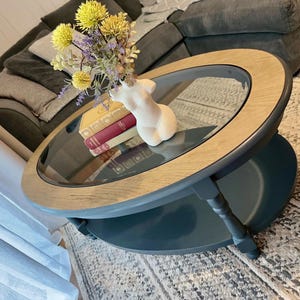 Può includere: Tavolino ovale con piano in vetro e bordo in legno chiaro. Il tavolo ha una base blu scuro e un ripiano inferiore. Un vaso in ceramica bianca a forma di torso contiene fiori e si trova sopra il tavolo, insieme a una pila di libri.