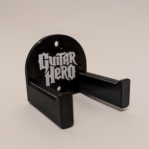 May include: A black wall-mounted controller holder featuring the Guitar Hero logo in white text on the front panel. The mount has a semi-circular back plate with two circular screw holes for installation and two protruding arms to hold a controller.
