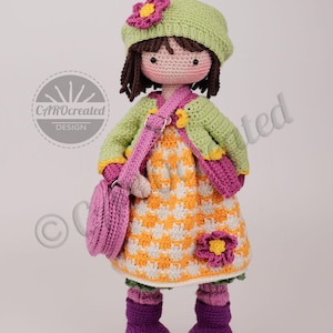 May include: A handmade crochet doll with a green hat and jacket, a yellow and white dress, and a pink purse. The doll has brown hair and is wearing purple boots. A pink flower is on the hat and dress. The doll is a CAROcreated DESIGN.