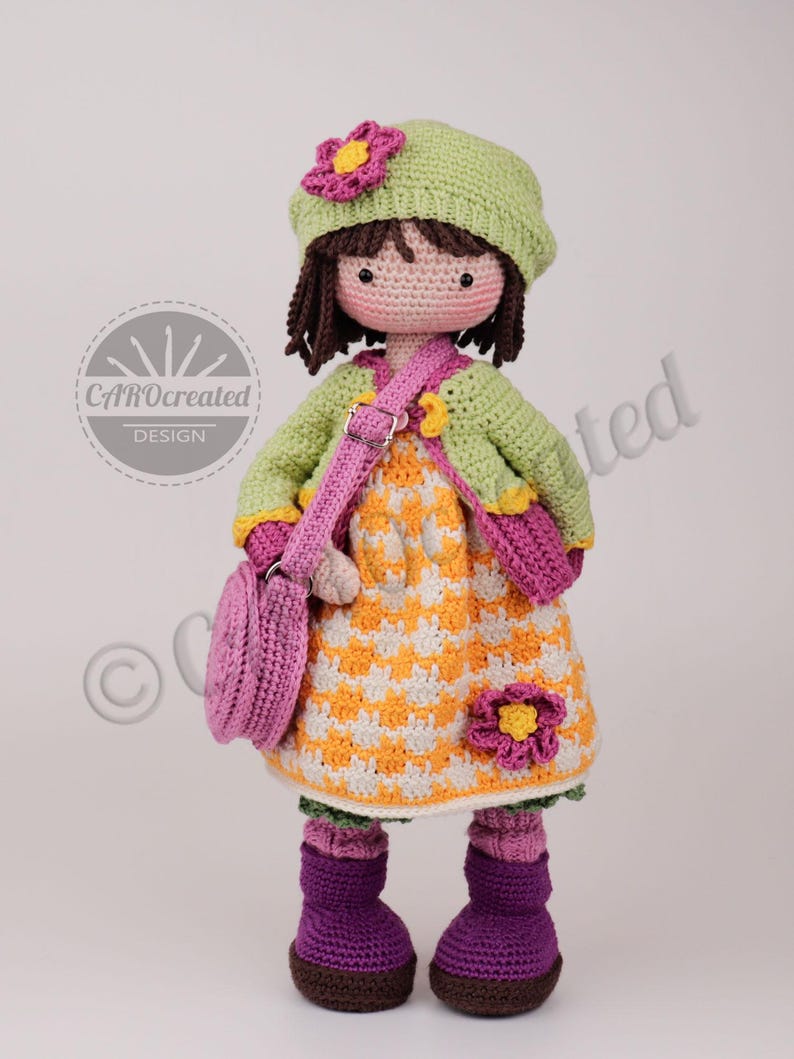 May include: A handmade crochet doll with a green hat and jacket, a yellow and white dress, and a pink purse. The doll has brown hair and is wearing purple boots. A pink flower is on the hat and dress. The doll is a CAROcreated DESIGN.