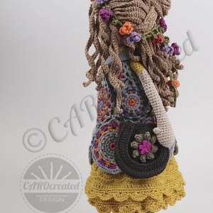 May include: A crocheted doll with long brown hair, wearing a colorful floral headband, a multi-colored crocheted vest, a yellow skirt with a crocheted lace trim, and brown shoes. The doll is holding a brown crocheted bag with a pink flower.