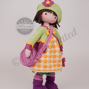 May include: A handmade crochet doll wearing a green hat with a pink flower, a green jacket, and a patterned dress. The doll has a pink purse and purple boots. The doll is standing and has brown hair and a smiling face.