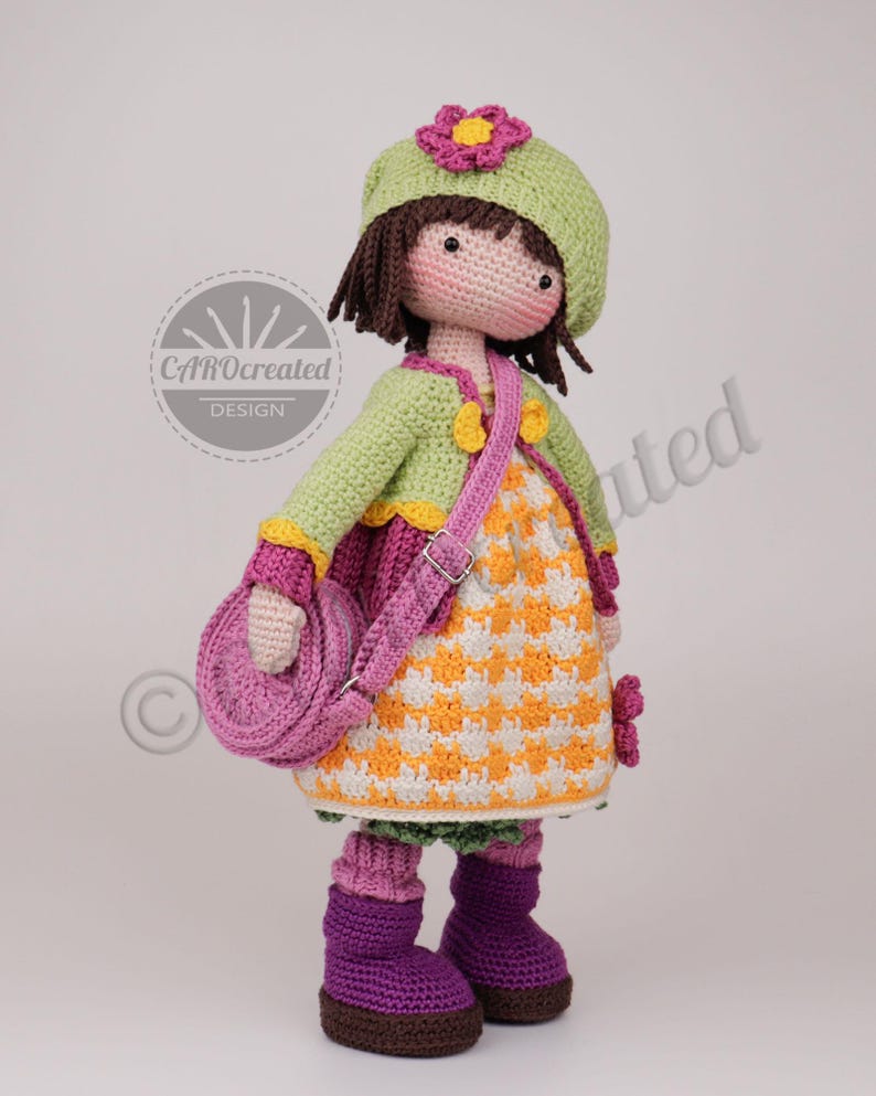 May include: A handmade crochet doll wearing a green hat with a pink flower, a green jacket, and a patterned dress. The doll has a pink purse and purple boots. The doll is standing and has brown hair and a smiling face.