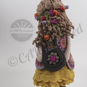 May include: Crocheted doll with brown hair, wearing a yellow skirt, a colorful vest, and a brown bag with a pink flower. The doll is holding a brown bag with a pink flower. The doll is standing on a white background.
