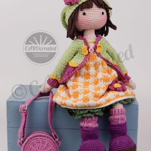 May include: A handmade crochet doll with brown hair, wearing a green jacket, orange and white dress, and purple boots. The doll has a matching green hat with a pink flower and carries a pink purse. The doll is sitting on a blue box.