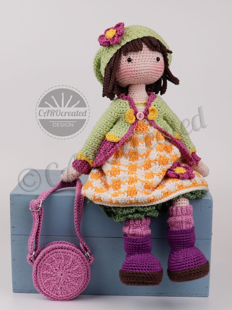 May include: A handmade crochet doll with brown hair, wearing a green jacket, orange and white dress, and purple boots. The doll has a matching green hat with a pink flower and carries a pink purse. The doll is sitting on a blue box.