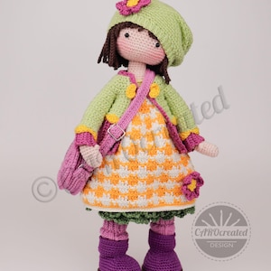 May include: A handmade crochet doll wearing a green hat, jacket, and a dress with an orange and white checkered pattern. The doll has a pink purse, purple boots, and a flower accent on the hat. The doll is a unique, handcrafted item.