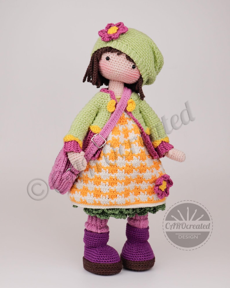 May include: A handmade crochet doll wearing a green hat, jacket, and a dress with an orange and white checkered pattern. The doll has a pink purse, purple boots, and a flower accent on the hat. The doll is a unique, handcrafted item.