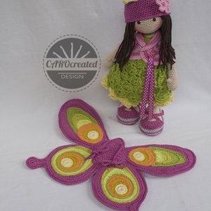May include: A crocheted doll with long brown hair wearing a green dress, pink vest, and pink hat with a flower. The doll is holding a pair of pink and green butterfly wings.