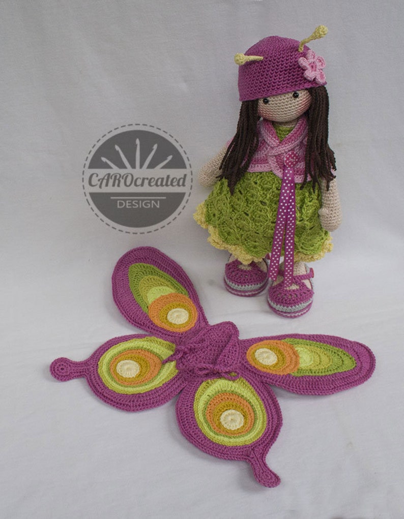 May include: A crocheted doll with long brown hair wearing a green dress, pink vest, and pink hat with a flower. The doll is holding a pair of pink and green butterfly wings.