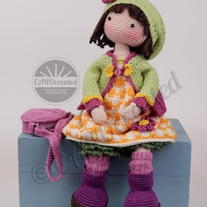 May include: A handmade crochet doll with a green hat and jacket, a yellow and white dress, and purple boots. The doll has a matching purple purse. The doll is sitting on a blue box. The doll's design is by CAROcreated DESIGN.