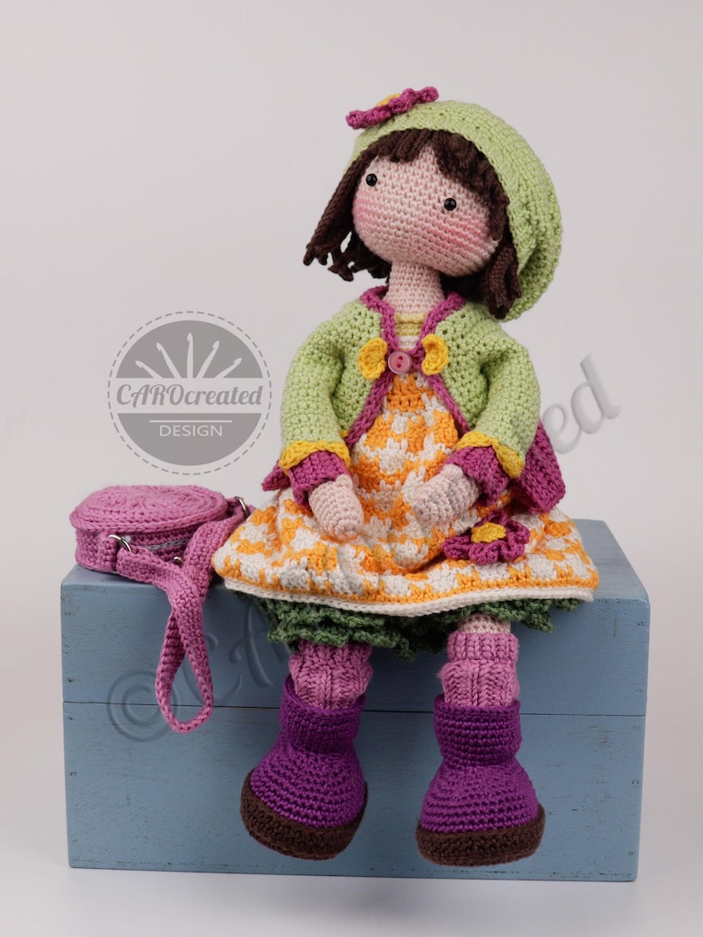 May include: A handmade crochet doll with a green hat and jacket, a yellow and white dress, and purple boots. The doll has a matching purple purse. The doll is sitting on a blue box. The doll's design is by CAROcreated DESIGN.