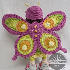 May include: A crocheted doll wearing a purple hat and butterfly wings with green, orange, and yellow accents. The doll is wearing a pink dress and shoes with buttons. The doll is standing on a white surface. The image includes the text "CAROcreated DESIGN".