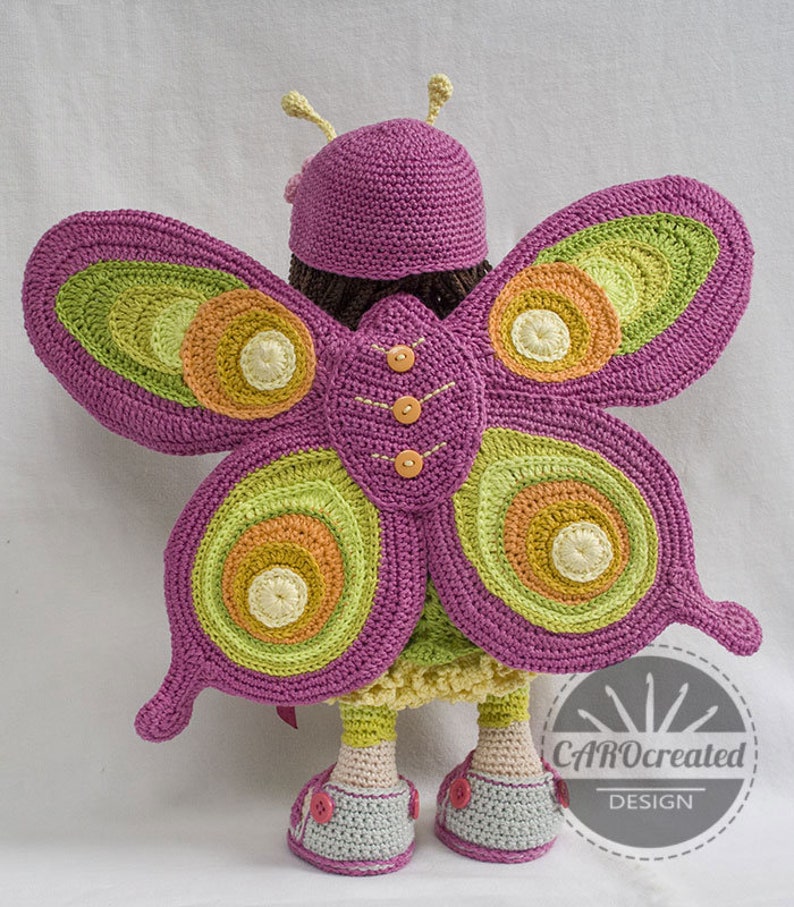 May include: A crocheted doll wearing a purple hat and butterfly wings with green, orange, and yellow accents. The doll is wearing a pink dress and shoes with buttons. The doll is standing on a white surface. The image includes the text "CAROcreated DESIGN".