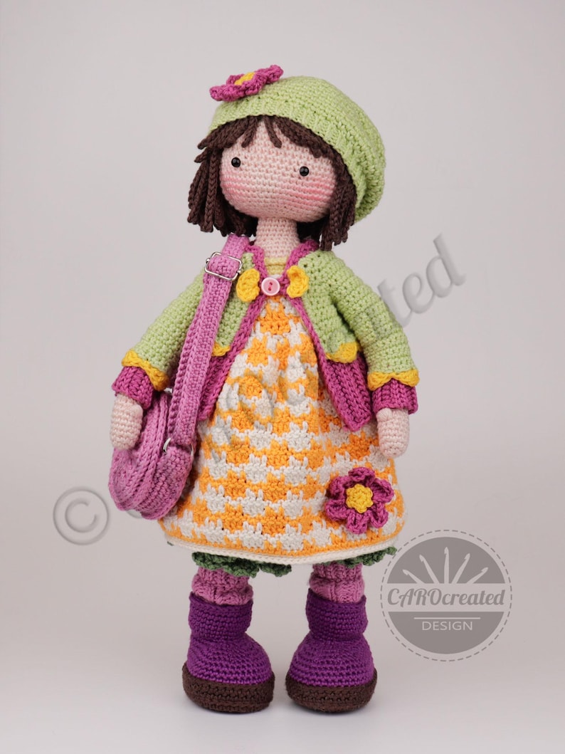 May include: A handmade crochet doll wearing a green hat, jacket, and purple boots. The doll has a yellow and white dress, a pink purse, and a flower accent. The doll is a charming, handcrafted item.