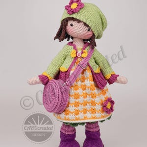 May include: A handmade crochet doll with a green hat, jacket, and dress. The doll has a pink purse, purple boots, and a flower on the hat. The dress is orange and white checkered. The doll is a whimsical, handcrafted item.
