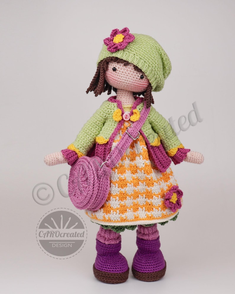 May include: A handmade crochet doll with a green hat, jacket, and dress. The doll has a pink purse, purple boots, and a flower on the hat. The dress is orange and white checkered. The doll is a whimsical, handcrafted item.