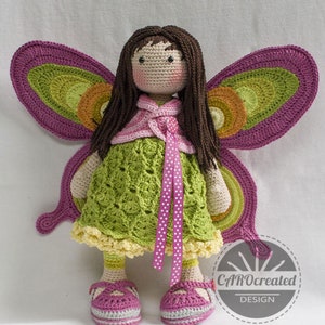 May include: A crocheted doll with brown hair and a green dress. The doll has butterfly wings made of crocheted yarn in shades of pink, green, and orange. The doll is wearing pink shoes and a pink ribbon around its waist. The doll is standing on a white surface. CAROcreated DESIGN is printed on a white circle in the lower right corner of the image.