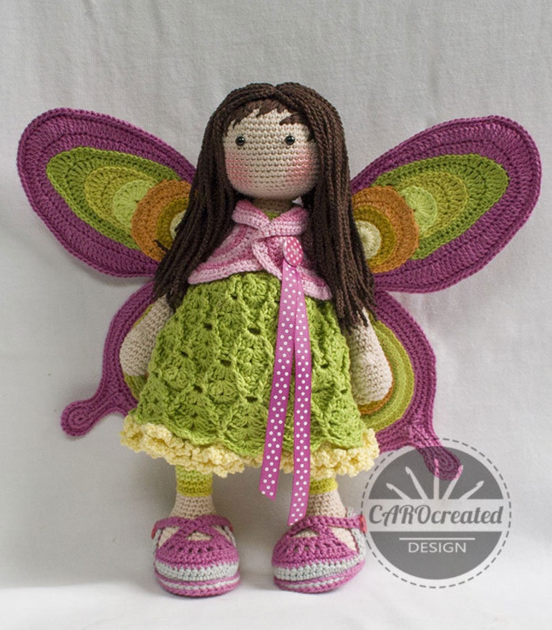 May include: A crocheted doll with brown hair and a green dress. The doll has butterfly wings made of crocheted yarn in shades of pink, green, and orange. The doll is wearing pink shoes and a pink ribbon around its waist. The doll is standing on a white surface. CAROcreated DESIGN is printed on a white circle in the lower right corner of the image.