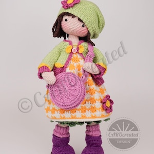 May include: A handmade crochet doll wearing a green hat with a pink flower, a green jacket, and a patterned dress. The doll has a pink purse and purple boots. The doll is approximately 12 inches tall.