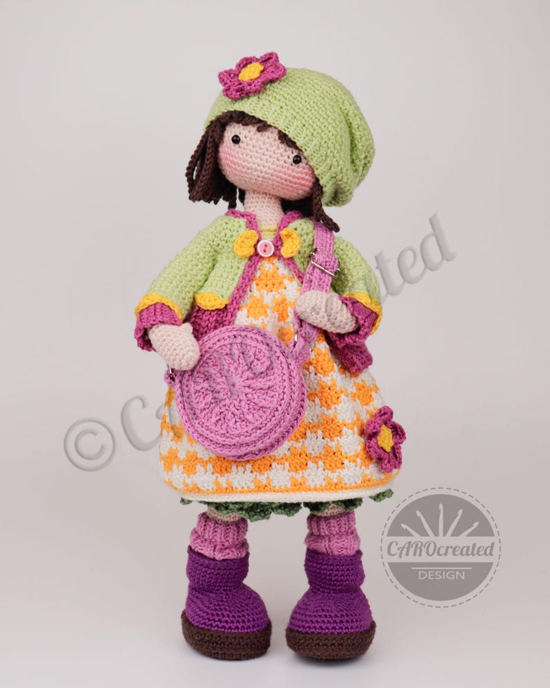 May include: A handmade crochet doll wearing a green hat with a pink flower, a green jacket, and a patterned dress. The doll has a pink purse and purple boots. The doll is approximately 12 inches tall.