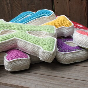 May include: A set of colorful crocheted letters, including pink, green, blue, yellow, and red, with white trim. The letters are arranged in a pile on a wooden surface.