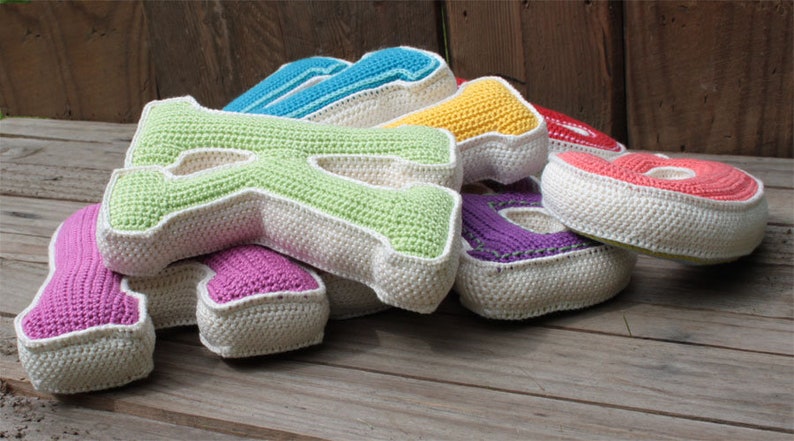 May include: A set of colorful crocheted letters, including pink, green, blue, yellow, and red, with white trim. The letters are arranged in a pile on a wooden surface.