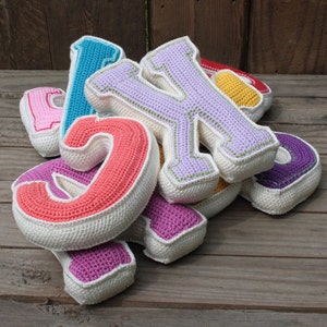 May include: A set of colorful crocheted letters, including pink, red, purple, blue, and yellow. The letters are made of yarn and have a white border. The letters are arranged in a pile on a wooden surface.
