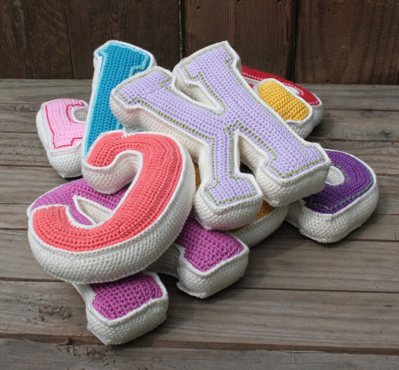 May include: A set of colorful crocheted letters, including pink, red, purple, blue, and yellow. The letters are made of yarn and have a white border. The letters are arranged in a pile on a wooden surface.