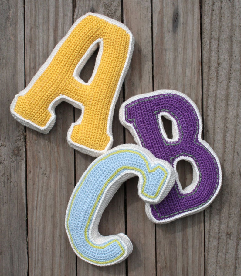 May include: Three crocheted letters, A, B, and C, are arranged on a wooden surface. The letter A is yellow, the letter B is purple, and the letter C is light blue.