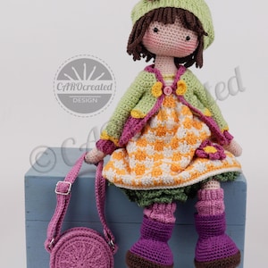 May include: A handmade crochet doll with a green jacket, orange and white dress, and purple boots. The doll has brown hair, a green hat with a flower, and a matching purple purse. The doll is sitting on a blue box.