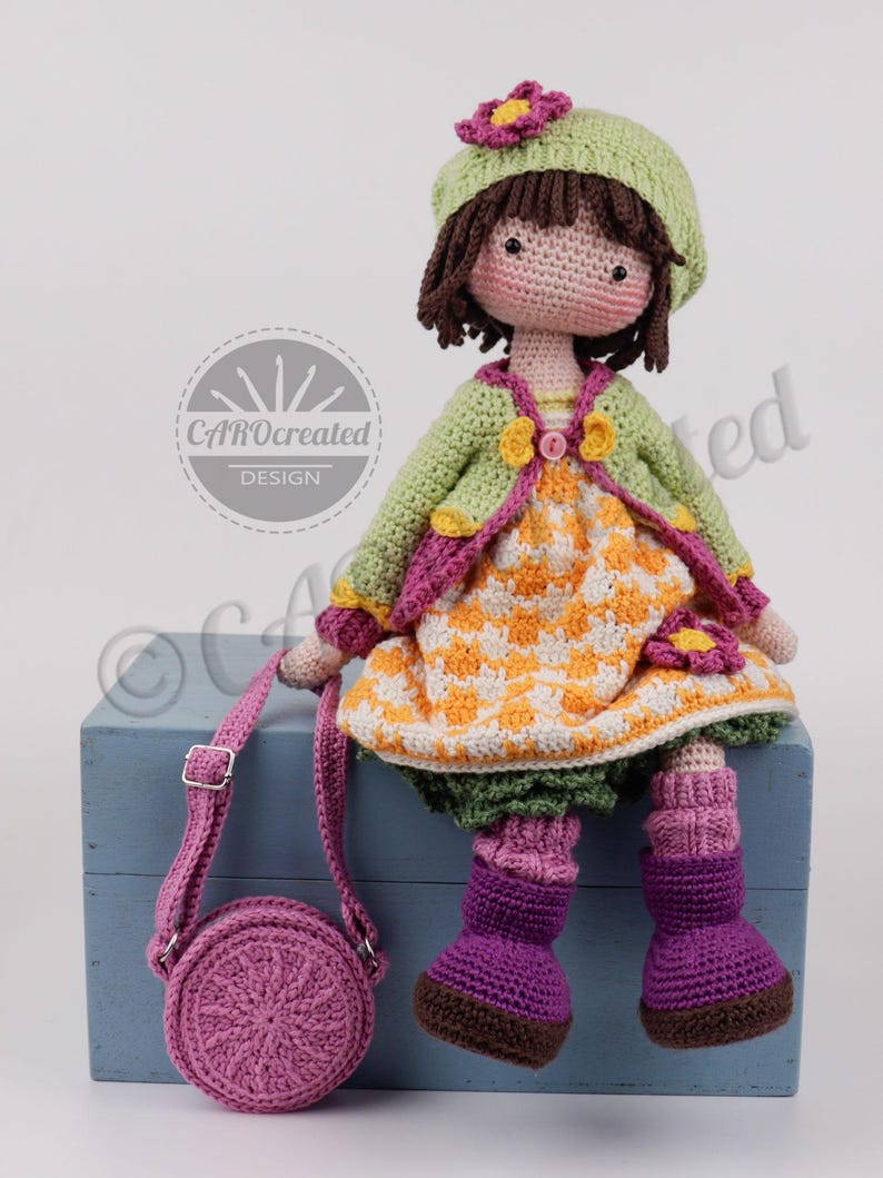 May include: A handmade crochet doll with a green jacket, orange and white dress, and purple boots. The doll has brown hair, a green hat with a flower, and a matching purple purse. The doll is sitting on a blue box.