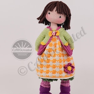 May include: A handmade crochet doll with brown hair and rosy cheeks. The doll wears a green jacket, an orange and white checkered dress, and purple boots. A pink flower is attached to the dress. The doll is a unique, handcrafted item.