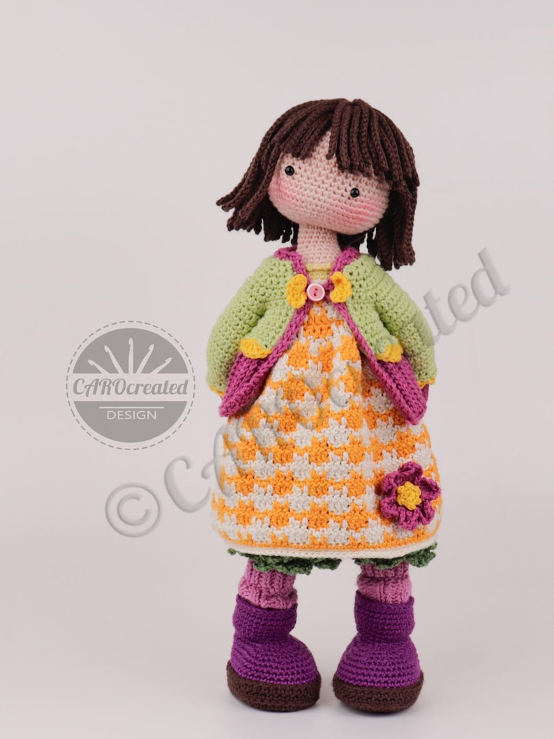 May include: A handmade crochet doll with brown hair and rosy cheeks. The doll wears a green jacket, an orange and white checkered dress, and purple boots. A pink flower is attached to the dress. The doll is a unique, handcrafted item.