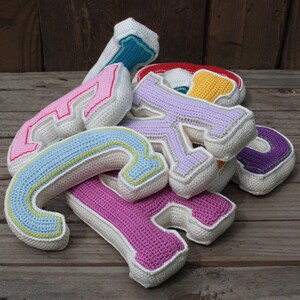 May include: A pile of crocheted letters in various colors, including pink, purple, blue, and yellow. The letters are made of white yarn with colored accents. The letters are arranged in a pile on a wooden surface.