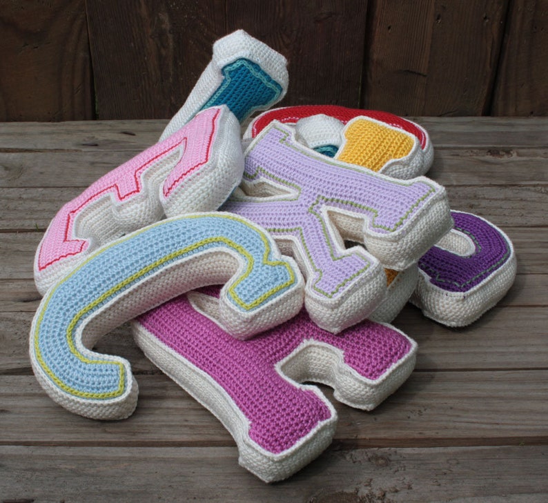 May include: A pile of crocheted letters in various colors, including pink, purple, blue, and yellow. The letters are made of white yarn with colored accents. The letters are arranged in a pile on a wooden surface.