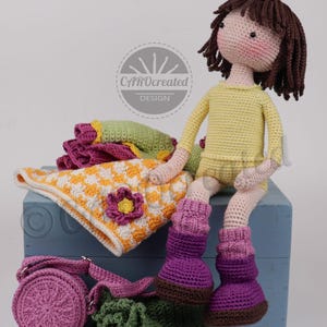 May include: A handmade crochet doll with brown hair, wearing a yellow outfit and purple boots, sits on a blue box. Accessories include a pink purse, a green bag, and a blanket with a flower design. The doll is surrounded by other crocheted items.