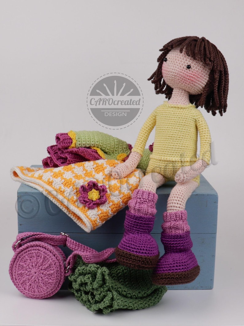 May include: A handmade crochet doll with brown hair, wearing a yellow outfit and purple boots, sits on a blue box. Accessories include a pink purse, a green bag, and a blanket with a flower design. The doll is surrounded by other crocheted items.
