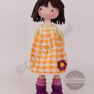 May include: A handmade crochet doll with brown hair and a light pink face. The doll wears a yellow and white checkered dress with a purple flower, purple boots, and pink leg warmers. The doll is approximately 12 inches tall.
