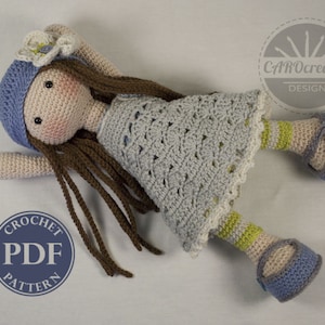 May include: Crochet doll pattern PDF. The doll is wearing a blue and white dress, a blue hat with a white flower, and blue shoes with a yellow stripe. The doll has brown hair and a sweet expression.