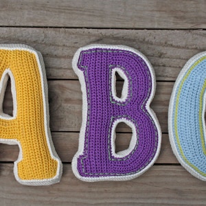 May include: Three crocheted letters, A, B, and C, are displayed on a wooden surface. The letter A is yellow, the letter B is purple, and the letter C is blue.