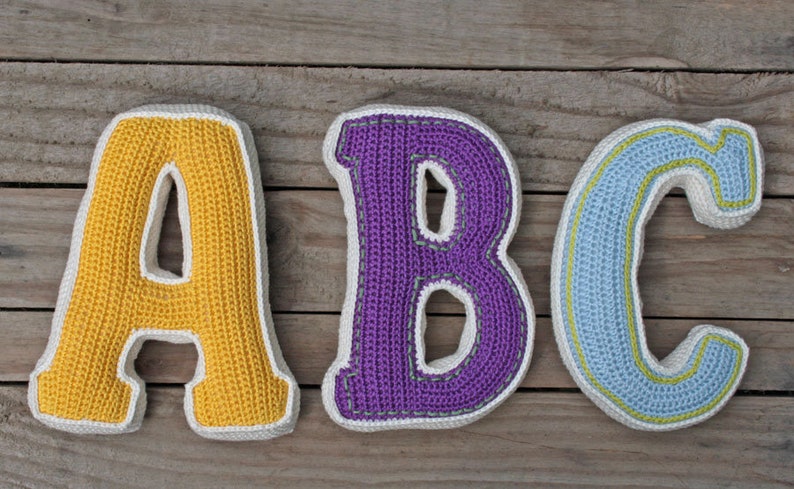 May include: Three crocheted letters, A, B, and C, are displayed on a wooden surface. The letter A is yellow, the letter B is purple, and the letter C is blue.